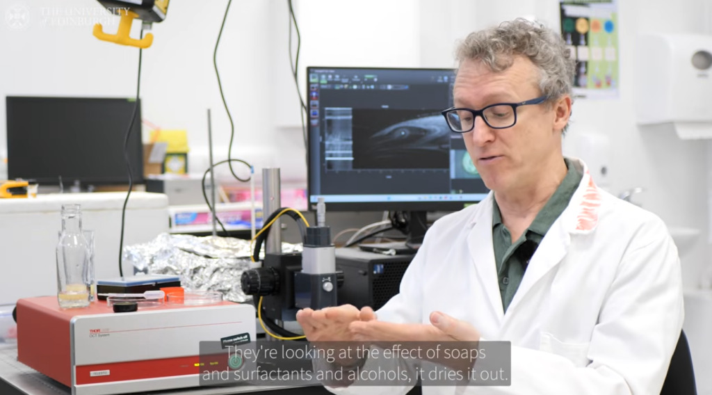 A man is wearing a white lab coat and is sitting in front of some laboratory equipment. He is explaining an idea using his hands.